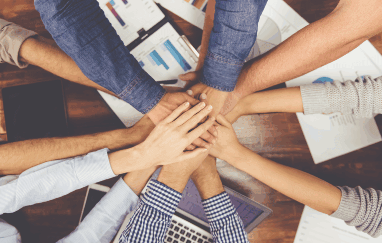 12 hands, joining "all-in" together over a table with business documents on it.